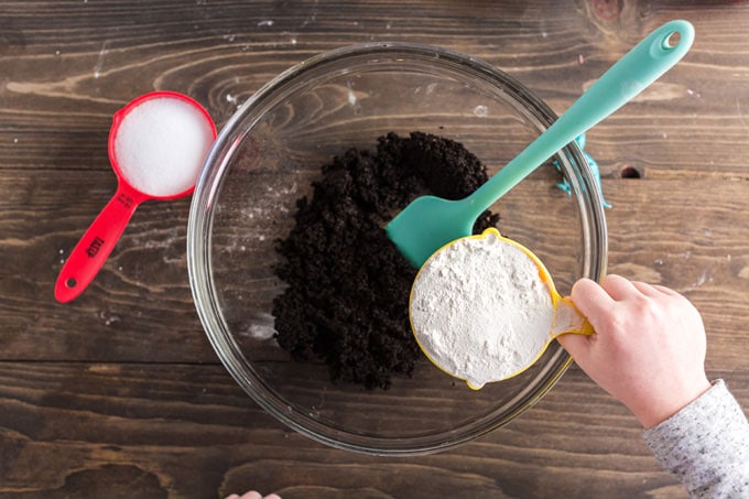 child holding measuring cup of flour over coffee grounds in a bowl