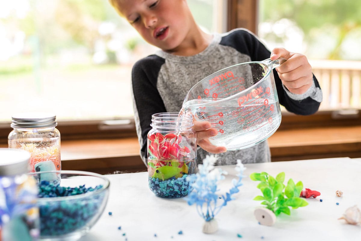 little boy adding water to a mason jar with aquarium plants and plastic fish